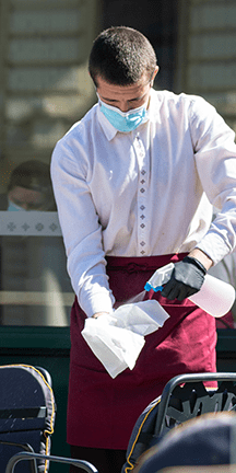Waiter wearing PPE spraying down cloth to wipe down tables and chairs